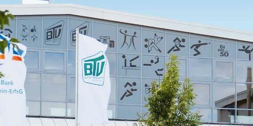 Fassade eines modernen Gebäudes mit großen Fenstern, Flaggen und sportlichen Symbolen. Grüne Pflanzen und Treppen im Vordergrund.