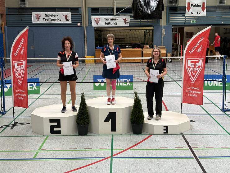 Platz 3 für Jutta Heimann-Feldhoff Drei Frauen stehen auf dem Siegertreppchen bei einem Badmintonturnier, jede hält eine Urkunde in der Hand.