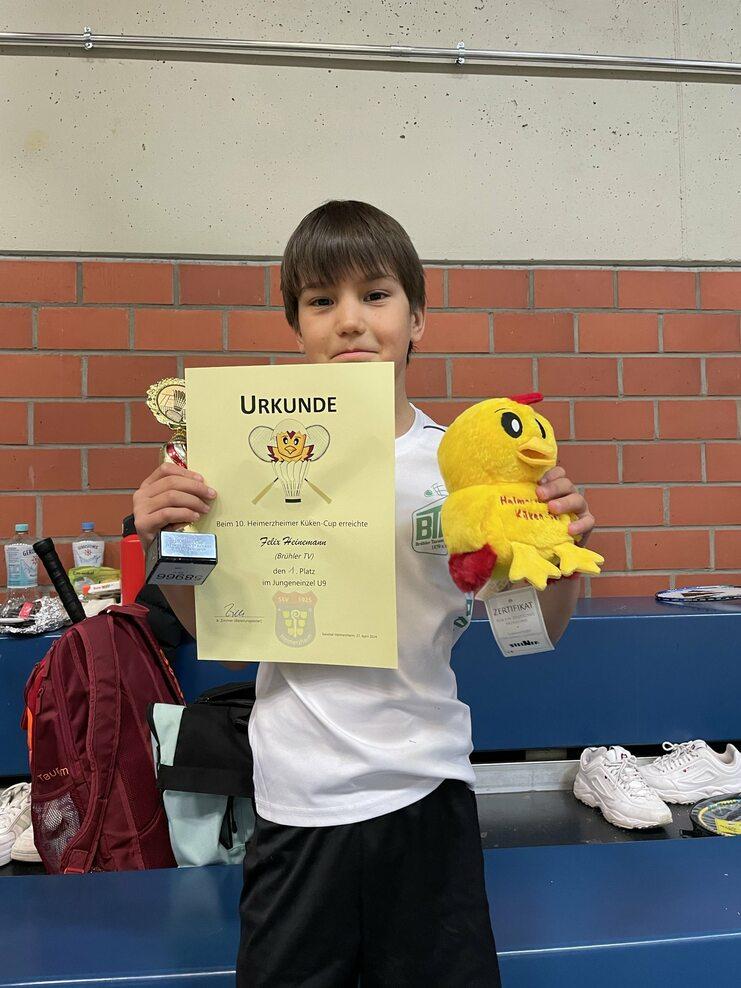 Felix gewinnt in Heimerzheim Ein Junge hält ein Diplom, einen Pokal und ein gelbes Stofftier in einer Sporthalle. Hintergrund mit Rucksack und Schuhen.