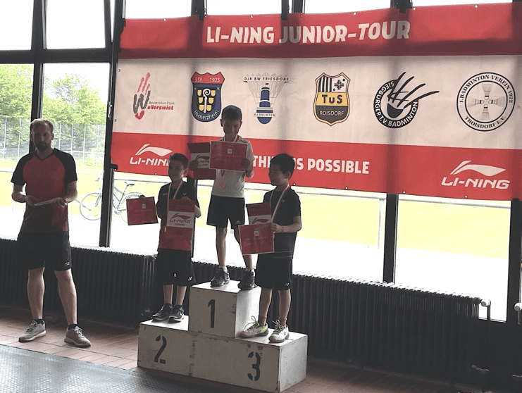 Drei Kinder mit Medaillen stehen auf dem Siegerpodest, während ein Trainer Urkunden überreicht. Banner im Hintergrund.