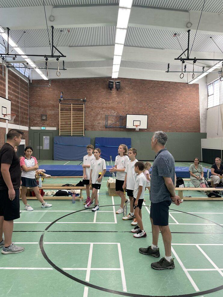 Gruppe von Kindern in Sportbekleidung hört einem Trainer in einer Turnhalle aufmerksam zu.