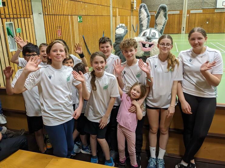 Gruppe von Jugendlichen in weißen T-Shirts, die fröhlich winken, während ein kostümiertes Kaninchen im Hintergrund steht.