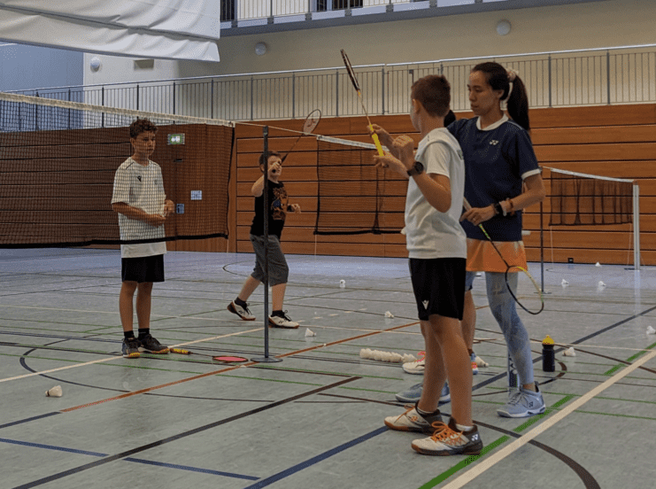 Vier Kinder mit Badmintonschlägern in einer Sporthalle, zwei besprechen etwas, während ein anderer spielt.