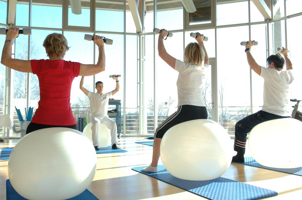Gruppe von vier Personen trainiert auf Gymnastikbällen mit Hanteln in einem hellen Fitnessraum.